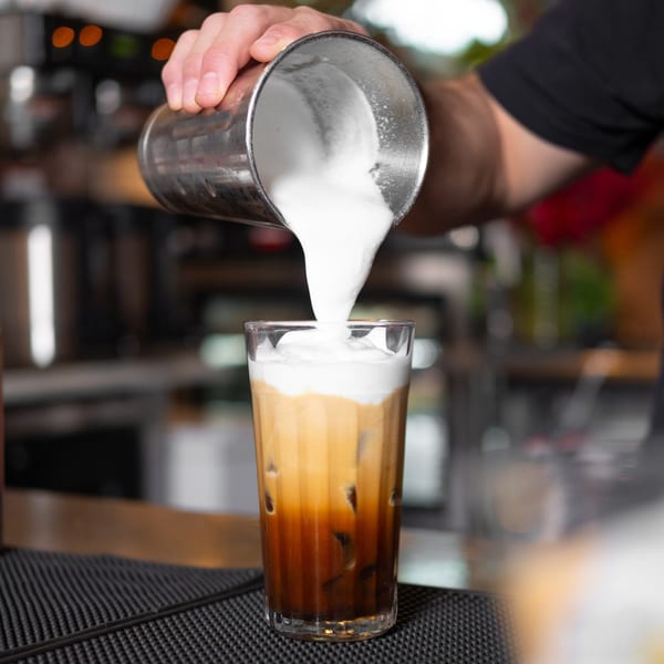 A barista pours foamed milk from a metal shaker into a tall glass of iced coffee.