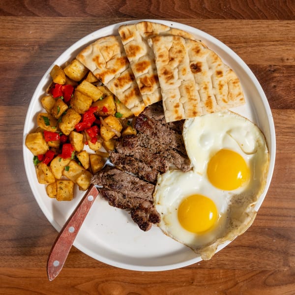 Steak and eggs breakfast with home fries and pita bread on a white plate.