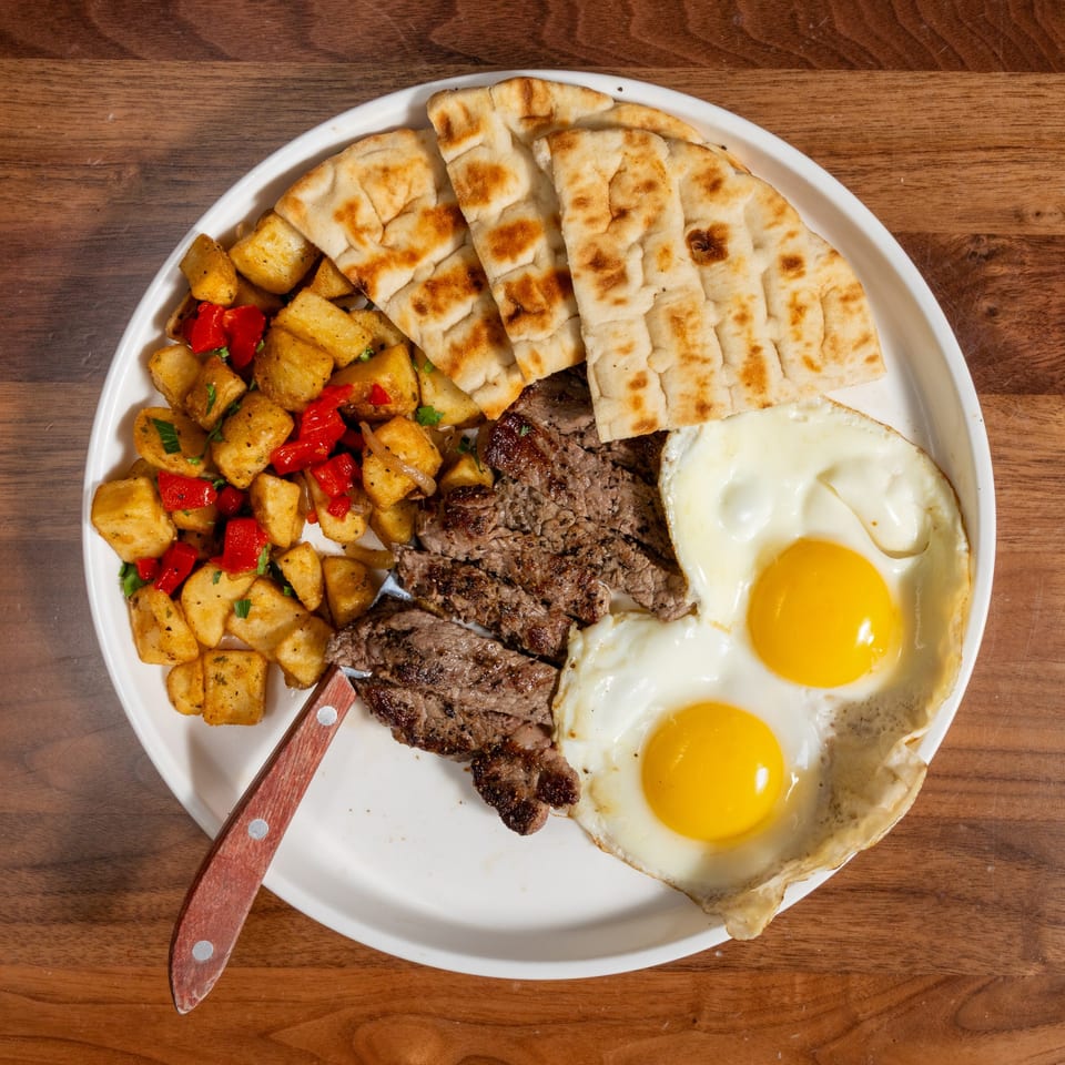 Steak and eggs breakfast with home fries and pita bread on a white plate.