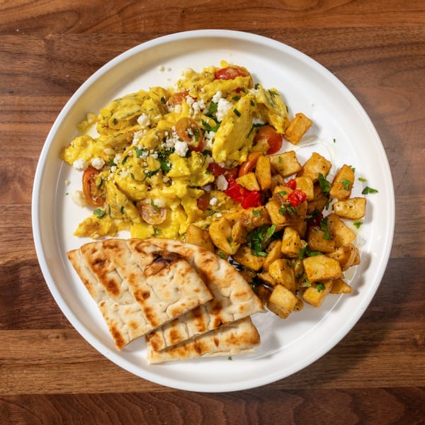 Scrambled eggs with feta and tomatoes, roasted potatoes, and pita bread on a white plate.