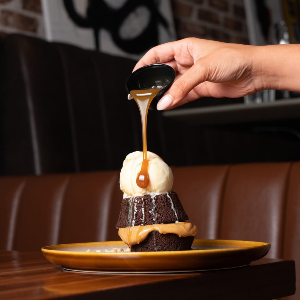 A hand pours caramel sauce over a chocolate lava cake with vanilla ice cream and peanut butter.