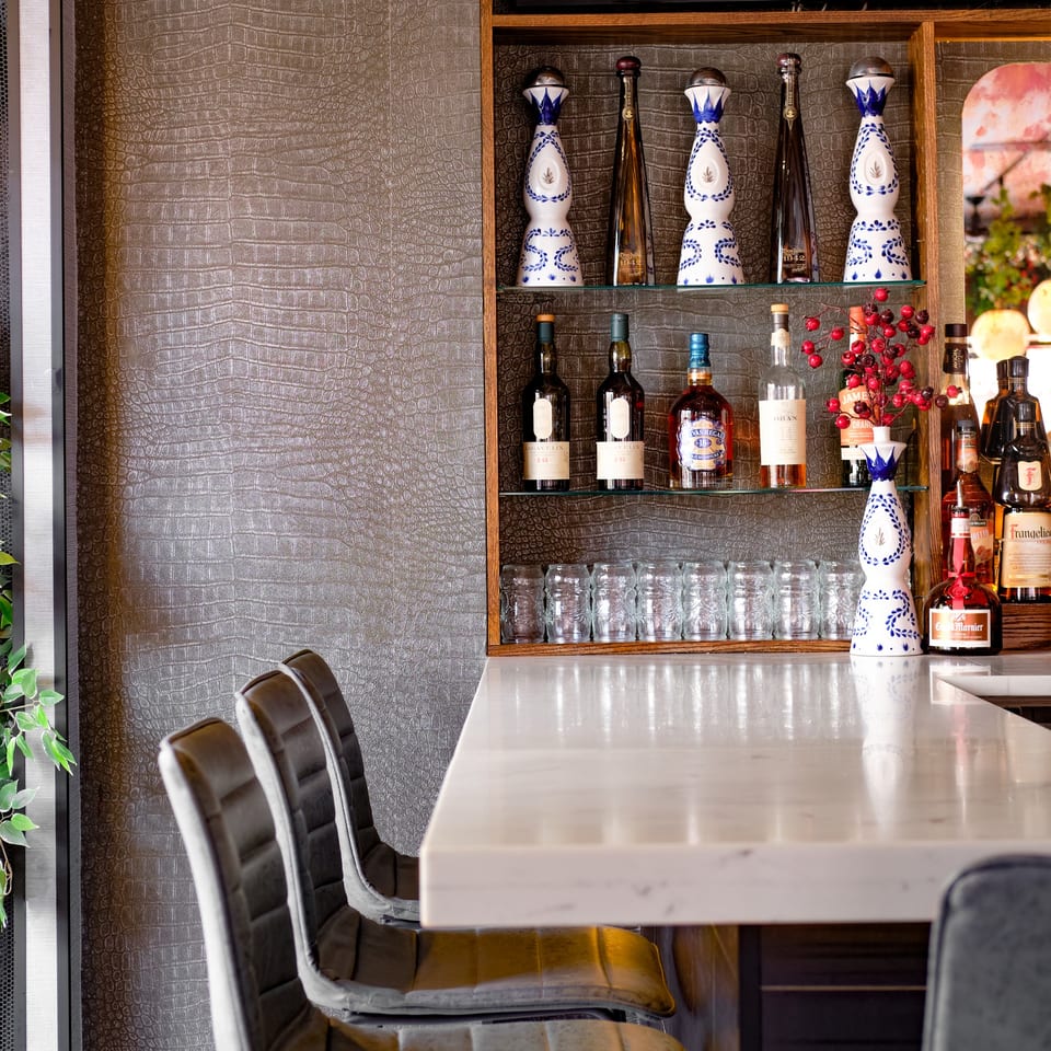 A bar with a white marble counter, bar stools, and shelves of liquor bottles and glasses.