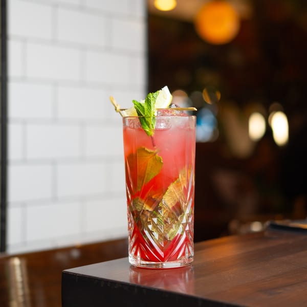A vibrant red brunch cocktail with mint and lemon garnish sits on a dark wooden bar.
