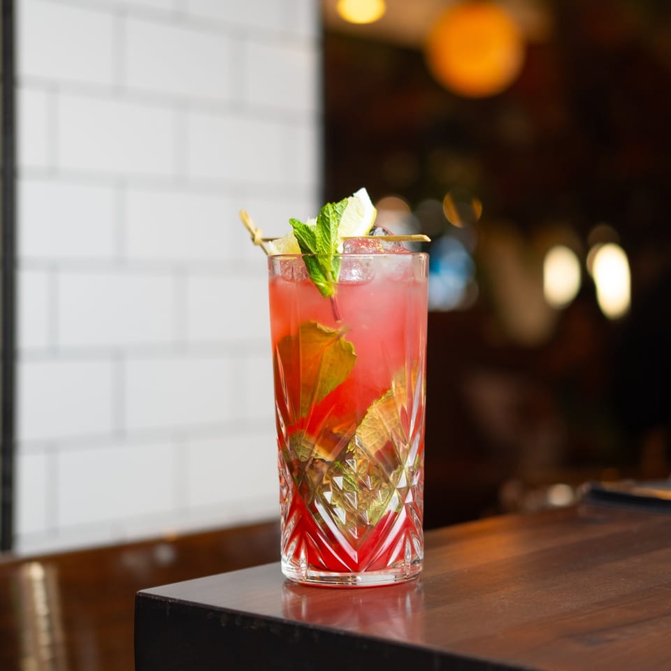 A vibrant red brunch cocktail with mint and lemon garnish sits on a dark wooden bar.