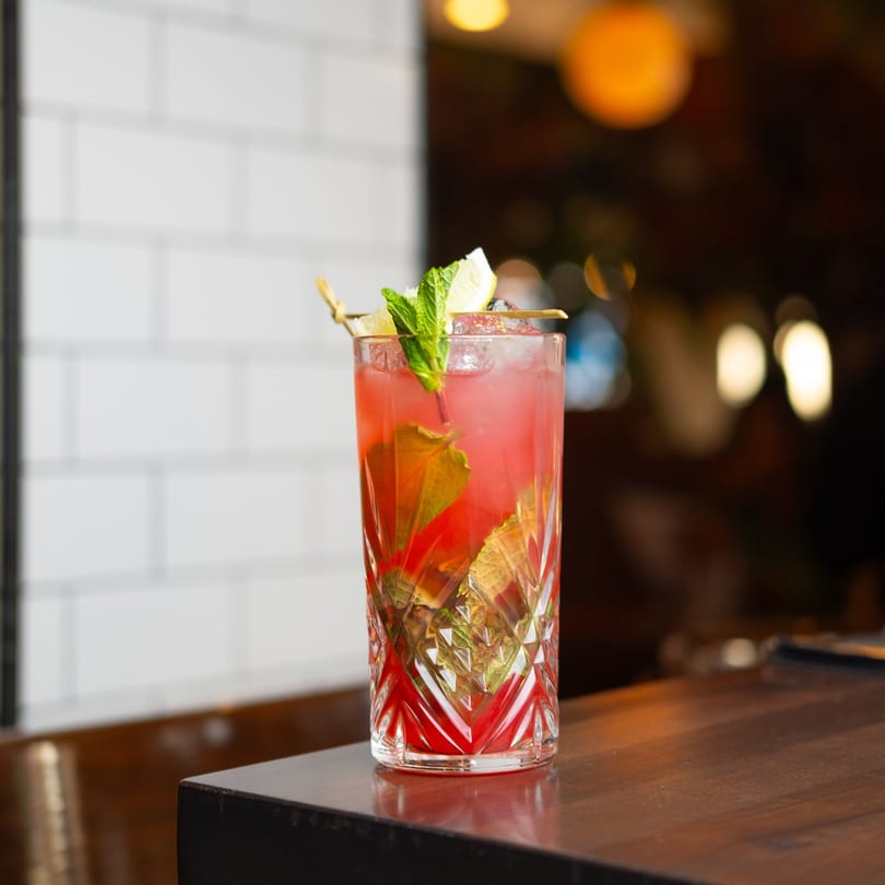 A vibrant red brunch cocktail with mint and lemon garnish sits on a dark wooden bar.