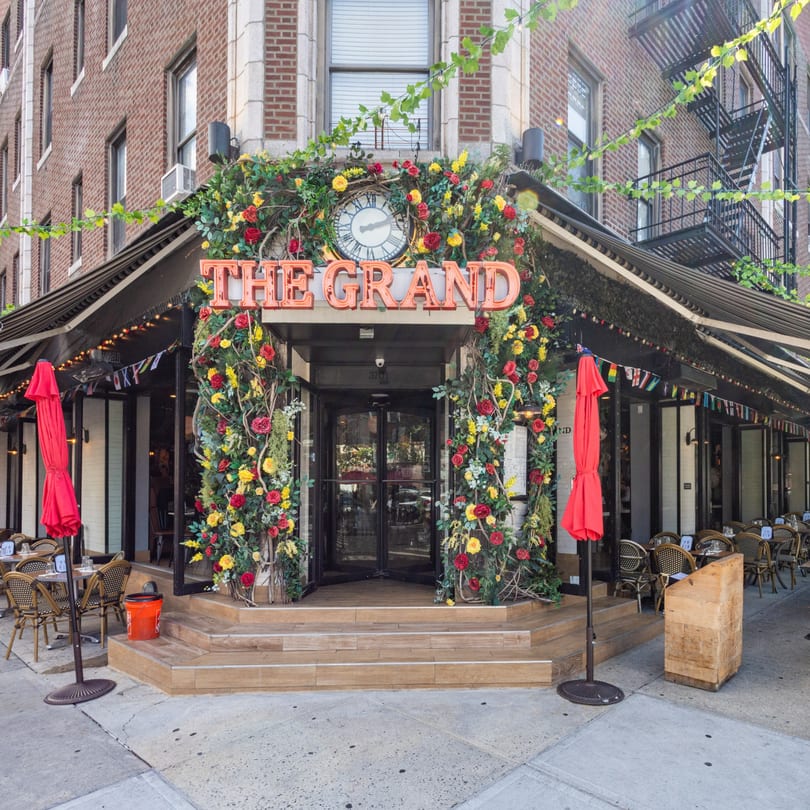 The Grand restaurant exterior with a floral archway, outdoor seating, and red umbrellas.