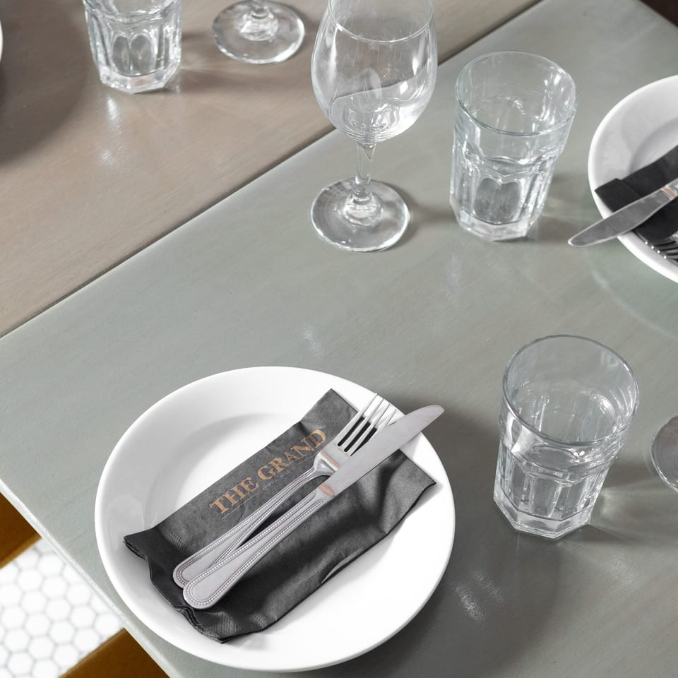 A set table at The Grand restaurant with a white plate, cutlery on a black napkin, and glasses.
