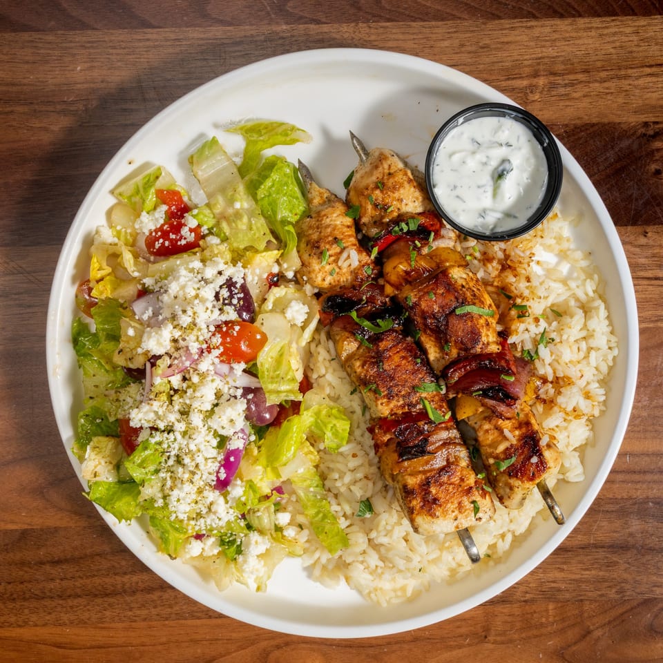 Overhead view of chicken skewers with rice, Greek salad, and tzatziki sauce on a white plate.