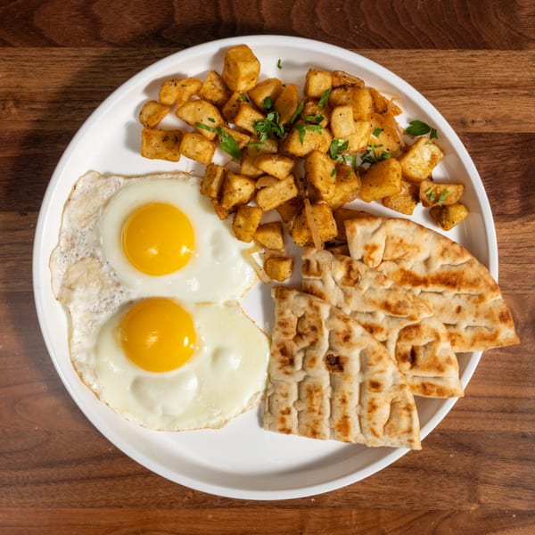 Breakfast plate with two fried eggs, seasoned potato cubes, and flatbread on a white plate.