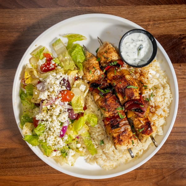 Grilled chicken skewers with rice, Greek salad, and a side of tzatziki sauce on a white plate.