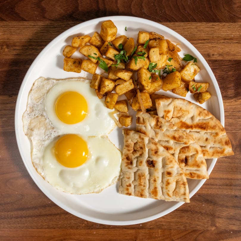 Breakfast plate with two fried eggs, seasoned potato cubes, and flatbread on a white plate.