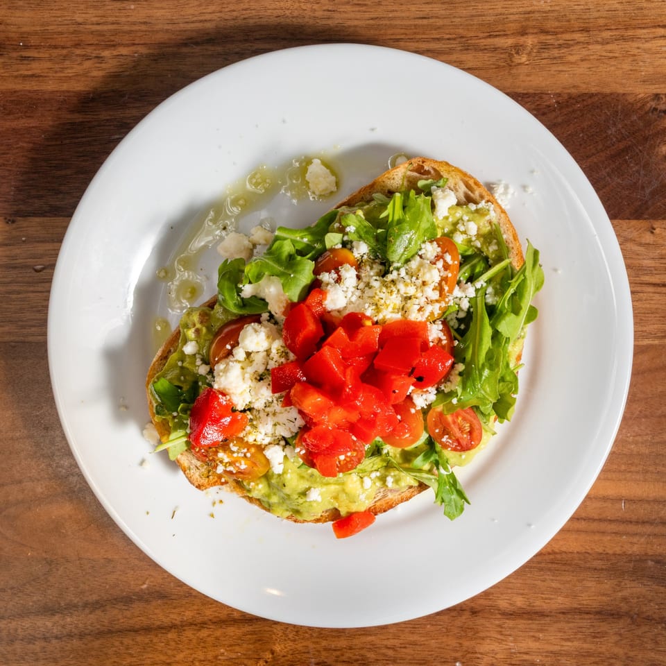 Avocado toast with arugula, diced tomatoes, feta cheese, and olive oil on a white plate.