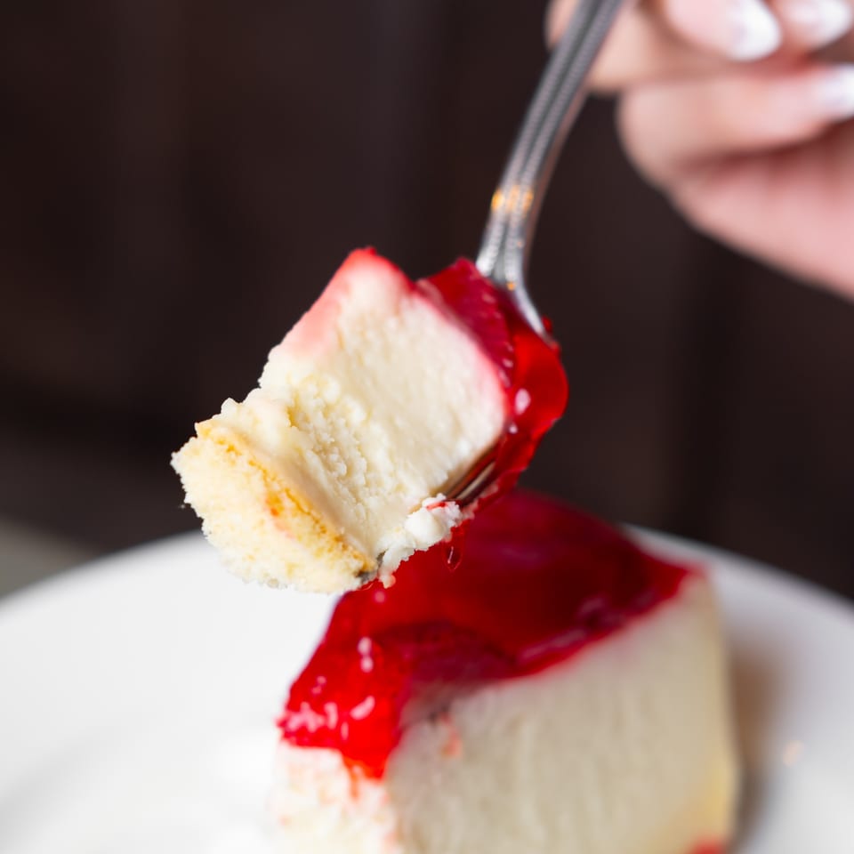 A fork lifts a bite of cheesecake with red fruit topping from a slice on a white plate.