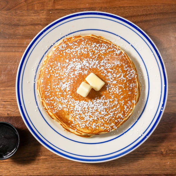 Stack of pancakes with powdered sugar and butter on a white plate with blue stripes, next to syrup.