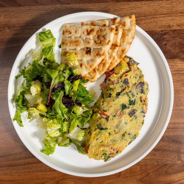 A white plate holds a savory omelet, fresh green salad, and three pieces of pita bread on a wooden table.