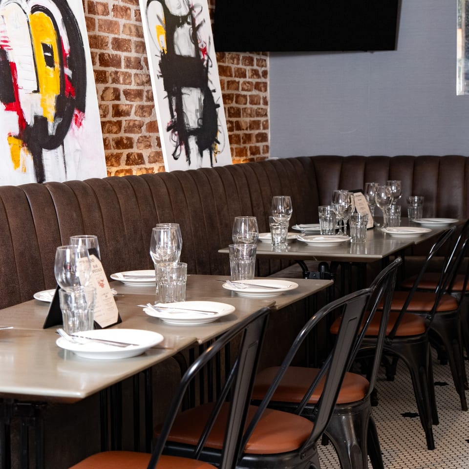 Restaurant interior with set tables, brown booth seating, brick wall, and modern art.