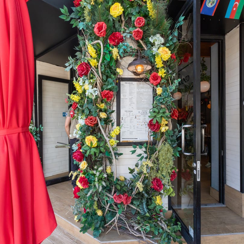 Restaurant entrance adorned with red and yellow flowers, green foliage, and a menu board.
