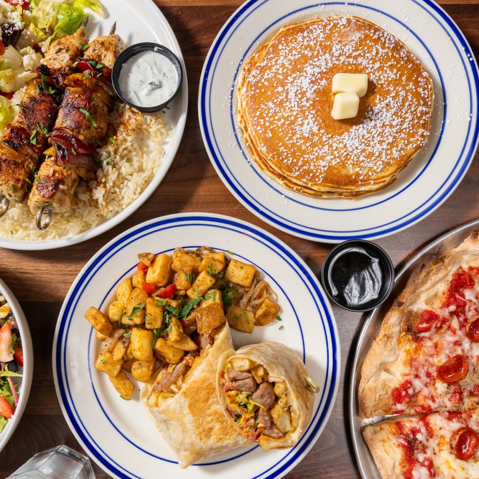 Overhead view of a table laden with diverse dishes: chicken skewers, pancakes, breakfast burrito, and pizza.