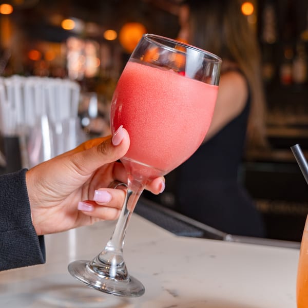 A hand holds a tall glass of a frosty pink drink, likely a frozen cocktail, at a bar.