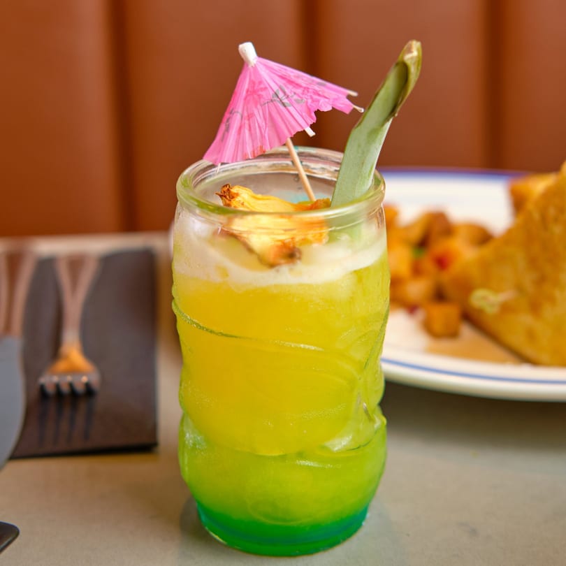 A vibrant green and yellow cocktail with a pink umbrella and pineapple garnish, next to brunch food.