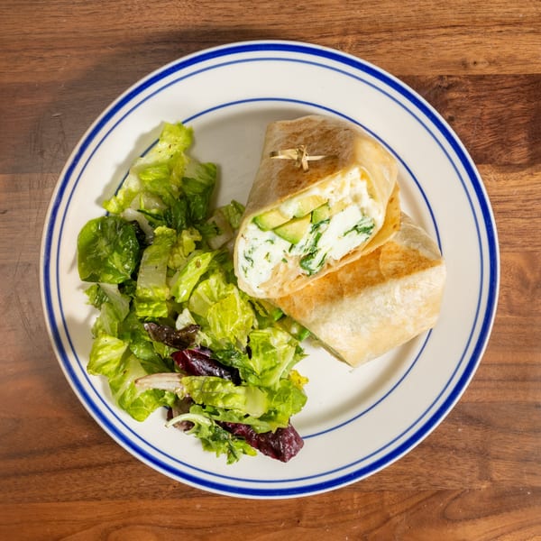 A white plate with blue stripes holds a cut wrap filled with avocado, cheese, and greens, alongside a fresh green salad.