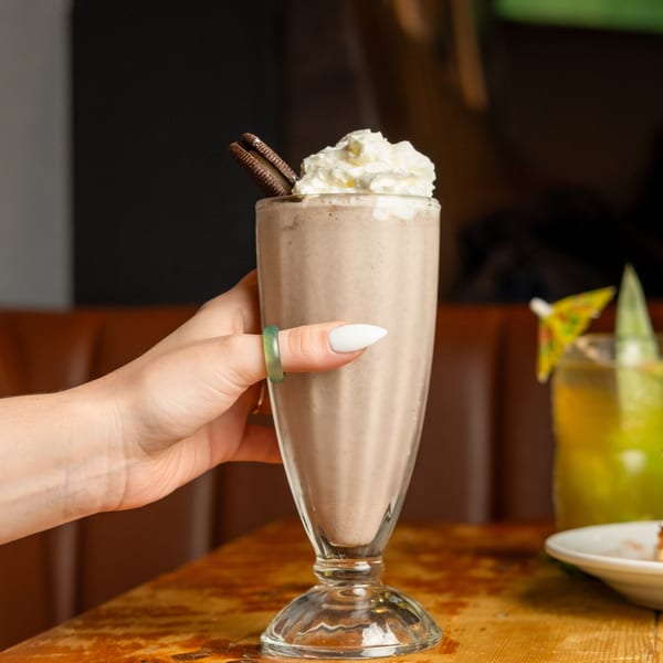 A hand holds a chocolate milkshake topped with whipped cream and two chocolate cookies.