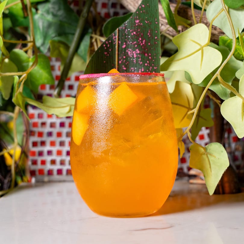 A vibrant orange cocktail with mango chunks and ice, garnished with a pink-dusted leaf.