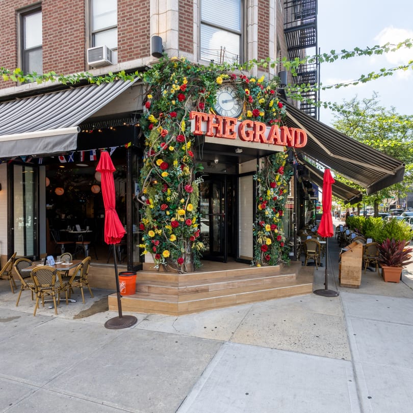 The Grand restaurant exterior with a floral archway, outdoor seating, and striped awnings on a sunny day.