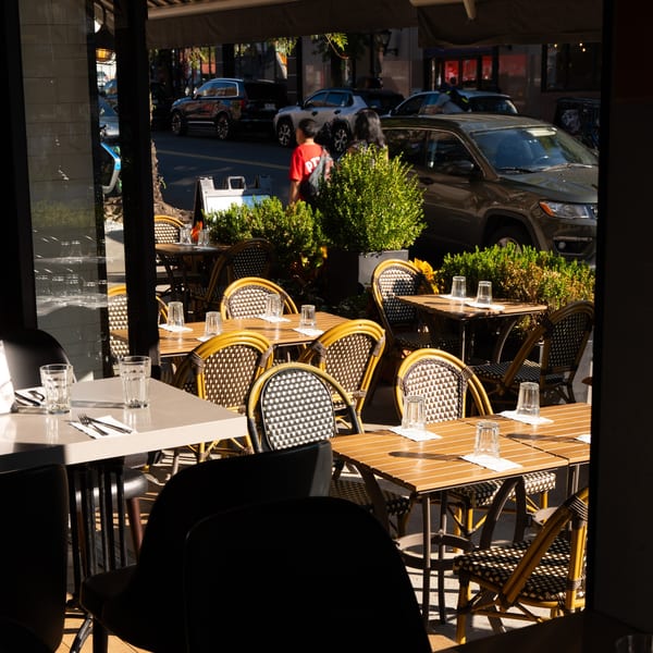 Outdoor dining area with empty tables and chairs, with a street and cars visible in the background.