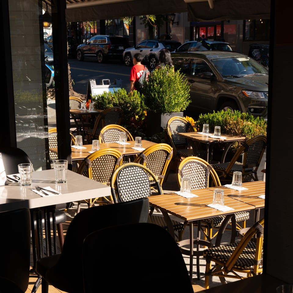 Outdoor dining area with empty tables and chairs, with a street and cars visible in the background.