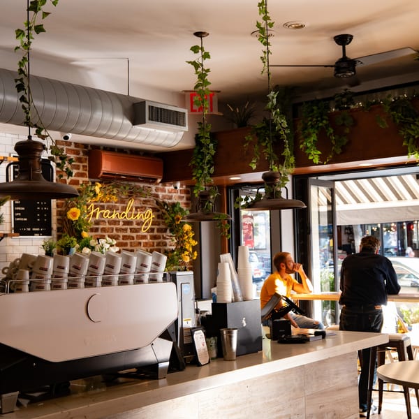 A coffee shop interior with a neon "Franding" sign, espresso machine, and two customers by the window.