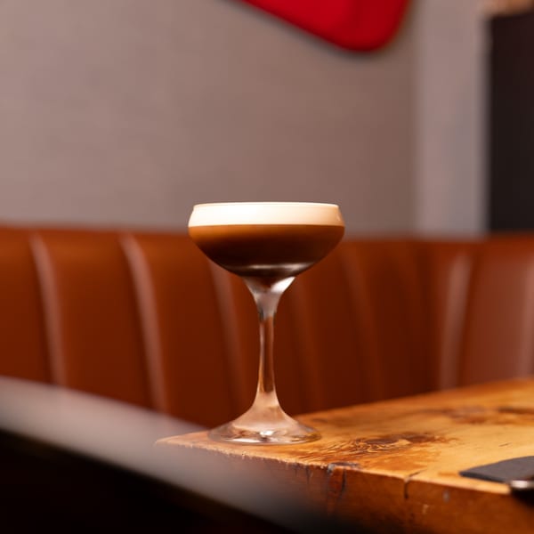 Espresso martini with a creamy foam top, on a wooden table with a brown leather booth.