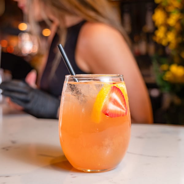 A refreshing orange cocktail with strawberry and orange slices, and a black straw, on a white bar.