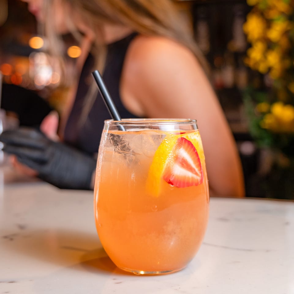 A refreshing orange cocktail with strawberry and orange slices, and a black straw, on a white bar.