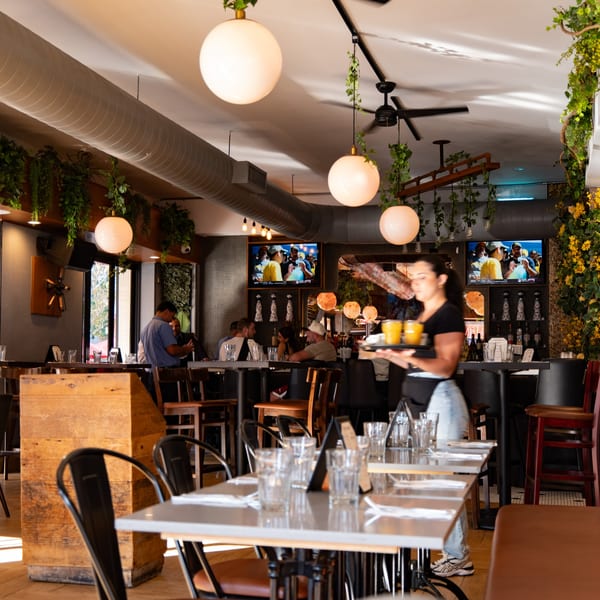 A restaurant interior with a server carrying drinks, patrons dining, and TVs showing sports.