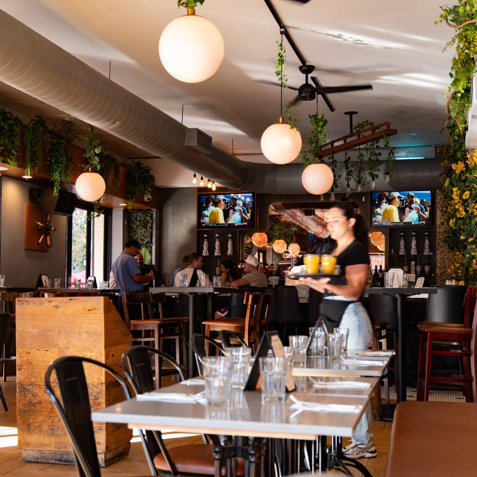 A restaurant interior with a server carrying drinks, patrons dining, and TVs showing sports.