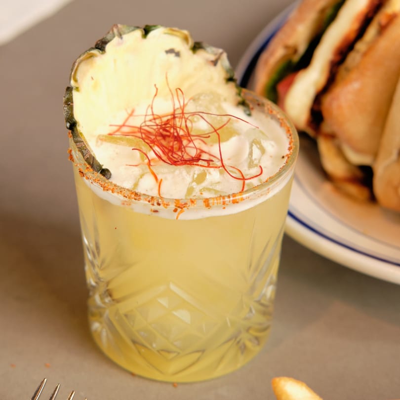 A pineapple cocktail with chili threads and a spiced rim, next to a sandwich and fries.