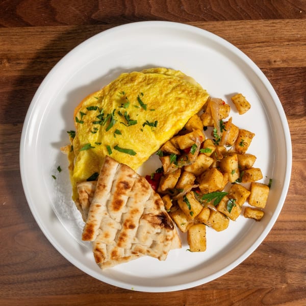 A fluffy omelet with parsley, crispy home fries, and warm pita bread on a white plate.